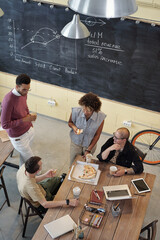 Poster - Group of young intercultural office workers having pizza at lunch break while standing and sitting by workplace