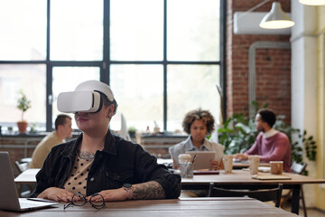 Poster - Young businesswoman in vr headset sitting by table in front of laptop and preparing presentation in working environment