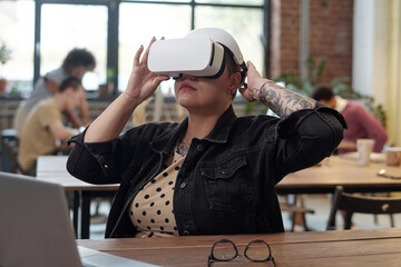 Poster - Young female office worker in vr headset watching virtual presentation while sitting by workplace in front of camera