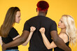 Love triangle. Relationship jealousy. Female competition. Multiracial group of  man embracing two fist threatening angry  women isolated on orange background.