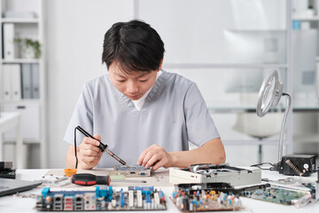 Poster - Young Chinese female technician in uniform repairing circuit board with microprocessor while sitting by workplace