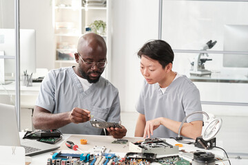 Poster - Young African man with circuit board consulting his female colleague while explaining how to fix tiny details or microchip