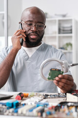 Poster - Young confident technician in uniform consulting client on mobile phone while looking at microchip through magnifying glass