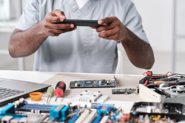 Poster - Hands of young African repairman photographing circuit board of electronic device while sitting by desk in workshop