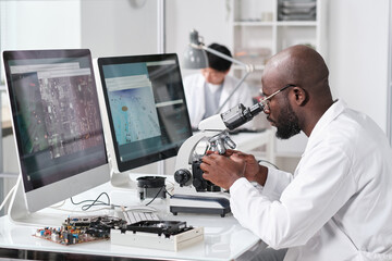 Poster - Young serious African man with microscope sitting by workplace in front of computer monitors and colleague on background
