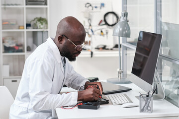 Poster - Serious young African repairman in whitecoat soldering microchip in circuit board while bending over desk in front of computer