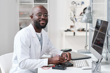 Poster - Young cheerful repairman or laboratory worker in whitecoat soldering details of circuit board by workplace and looking at you