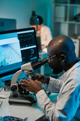 Poster - Young man of African ethnicity sitting by workplace and looking in microscope against computer screen and colleague