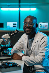 Poster - Happy young male laboratory worker in whitecoat and eyeglasses sitting by desk with microscope and looking at you with smile