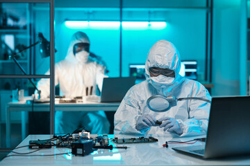 Poster - Young female scientist in protective workwear looking at microchip through magnifying glass against colleague in laboratory