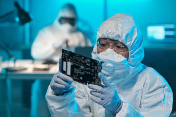 Poster - Young Asian female in protective workwear looking at circuit board in her gloved hands while working in laboratory