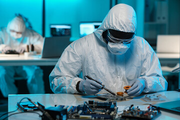 Poster - Contemporary technician in protective workwear soldering tiny parts of circuit board while sitting by desk in laboratory