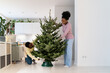Family Christmas tradition. Young african american woman helping husband to put freshly cut live xmas tree straight in stand, enjoying New Year preparation together, decorating home for festive season