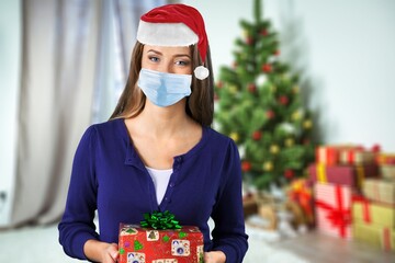 Wall Mural - Portrait of a beautiful girl in medical mask and santa hat with a present in hands, standing in front of decorated christmas tree at home