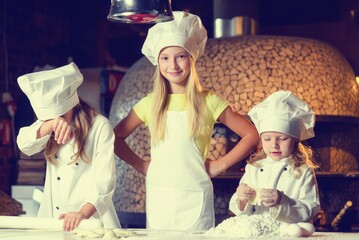 Near the oven, three cheerful little girls, soiled in flour and dressed in chef's uniforms, are preparing a flour product.