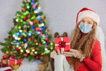 Wall Mural - Portrait of a beautiful girl in medical mask and santa hat with a present in hands, standing in front of decorated christmas tree at home