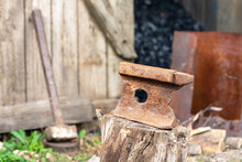 Old Rusty Anvil Stands On A Stump In The Backyard Of Village House, Sledgehammer Leaning On Wooden Fence Enclosing A Barn With Coal On Blurred Background. Heavy Working Tools.