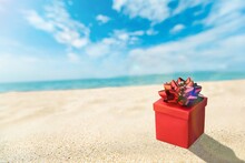 Beautiful Gift Box With A Ribbon Standing On The Sand With Blue Sea And Sun On Background.