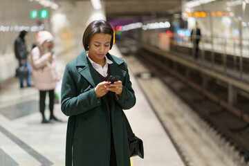 Young businesswoman waiting on subway platform for carriage reading text or business email on smartphone. Busy african woman with mobile phone chatting with colleague while returning home from office