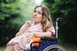 Portrait of young caucasian woman with disability in stylish summer dress looking at camera with green bushes on background. Time spending in fresh air. Lifestyles of wheelchair user.