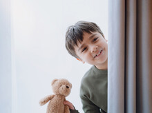 Cute Young Boy Holding Teddy Bear Standing Behind Lace Curtain With Bright Light In Morning, Happy Kid With Smiling Face On Window Sill And Plays At Hide-and-seek Behind Curtain