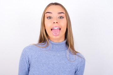 Young caucasian girl wearing blue turtleneck over white background with happy and funny face smiling and showing tongue.