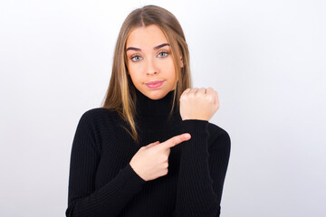 Young caucasian girl wearing black turtleneck over white background in hurry pointing to watch time, impatience, upset and angry for deadline delay