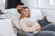 © Andrii - Happy family concept. Husband hug belly pregnant wife sitting floor indoor living room near sofa