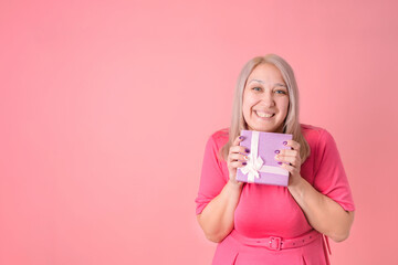 A joyful 40-year-old woman hugs a gift box standing on a pink background. Copyspace