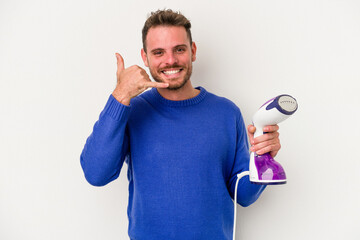 Young caucasian man holding an iron isolated on white background showing a mobile phone call gesture with fingers.