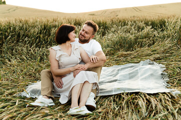 beautiful middle-aged couple in a wheat field sitting on the bedspread and hugging tenderly at sunset. older people in love. lovers having a picnic in the field