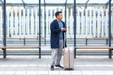 Asian businessman tourist, waiting for a taxi near the airport, uses the phone app to rent a car, a man with a big suitcase