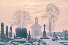 Winter Graveyard At Christmas With Sunny Clouds Over The Cross And Morgue. Snowy And Foggy Cemetery  With Two Linden Trees And Soft Fog Above Graves With Gravestones.