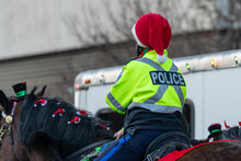 A Female Police Officer Rides On The Back Of A Large Black Police Mounted Unit Horse During A Christmas Parade. The Animal Has Decorations In Its Mane. The Cop Is Wearing A Red Christmas Hat. 