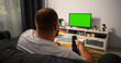 © RecCameraStock - Back view Man sitting on the couch in the living room Watching TV with Green Screen and Changing Channels with Remote Control in his hand. Chroma Key TV in focus.