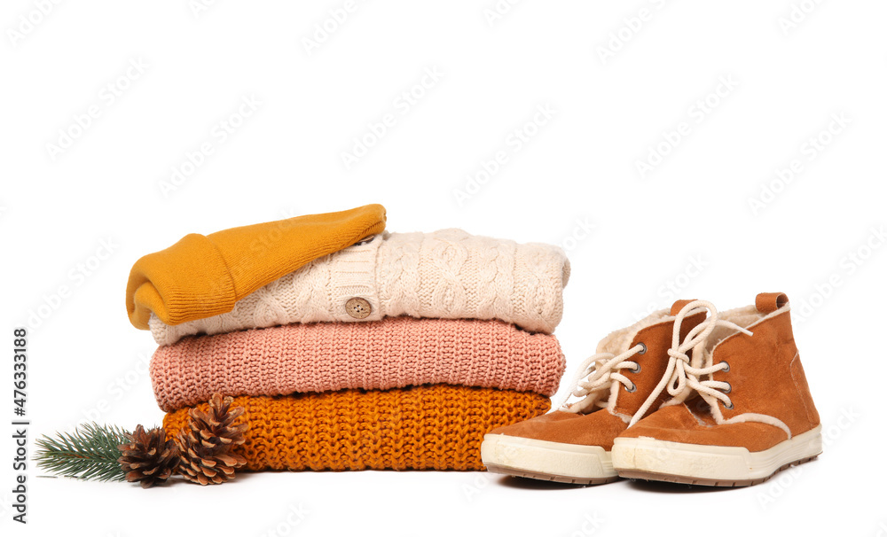 Stack of knitted sweaters, shoes, hat and winter decor on white background