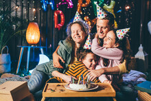 Cheerful Father Hugs Curly Haired Wife And Cute Children With Party Hats Near Small Table With Cake At Birthday Celebration In Cottage Yard In Evening
