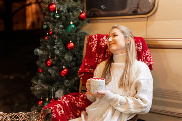 Wall Mural - young candid woman in knitted cozy sweater holding in hands cup of cocoa drink with marshmallow sitting relaxing on chair camping outdoor and enjoyment happy new year and merry christmas holidays eve