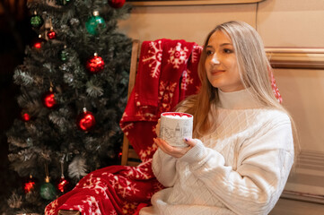 Wall Mural - young candid woman in knitted cozy sweater holding in hands cup of cocoa drink with marshmallow sitting relaxing on chair camping outdoor and enjoyment happy new year and merry christmas holidays eve