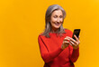 © Vadim Pastuh - Cheerful attractive senior woman holding smartphone, enjoying chatting in social networks, texting messages with congratulations, smiling, using mobile app, isolated on yellow