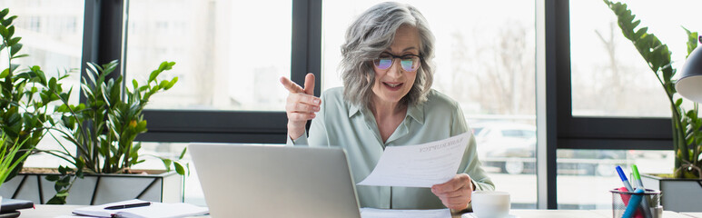 Wall Mural - Mature businesswoman holding paper during video call on laptop, banner.