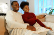 © DimaBerlin - Movie time. Young happy african american family couple man and woman enjoy weekend together at home, watch comedy show on tv and laughing, happy black husband and wife with remote controller on couch