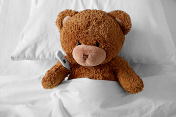 Wall Mural - medicine, healthcare and childhood concept - ill teddy bear toy head with thermometer lying in bed