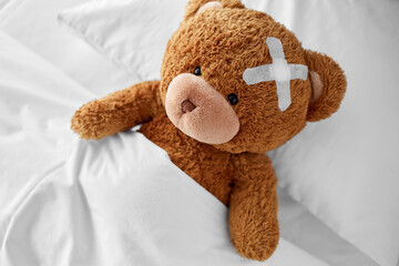 Wall Mural - medicine, healthcare and childhood concept - ill teddy bear toy with medical patch on head lying in bed