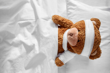 Wall Mural - medicine, healthcare and childhood concept - ill teddy bear toy with bandaged paw and head lying in bed