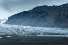 People In Red And Blue Outdoor Jackets Dwarfed By A Glacier On A Cold, Cloudy Day In Southern Iceland.