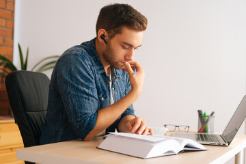 Wall Mural - Medium shot of concentrated pensive young man reading business paper book sitting at desk with laptop at home office. Focused student or freelancer male learning alone with textbook in light room.