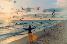 Seascape During Sunrise With Beautiful Sky. Young Happy Woman With Her Hands In The Air Walks Along The Beach In An Orange Dress. Seagulls Fly Over The Beach