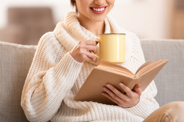 Wall Mural - Young woman drinking tasty tea and reading book on sofa at home