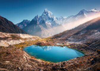 Wall Mural - Snowy mountain with illuminated peaks and small lake with blue water at sunrise in Nepal. Beautiful landscape with lake, high rocks in snow, golden sun rays, hills at dawn. Himalayan mountains. Nature
